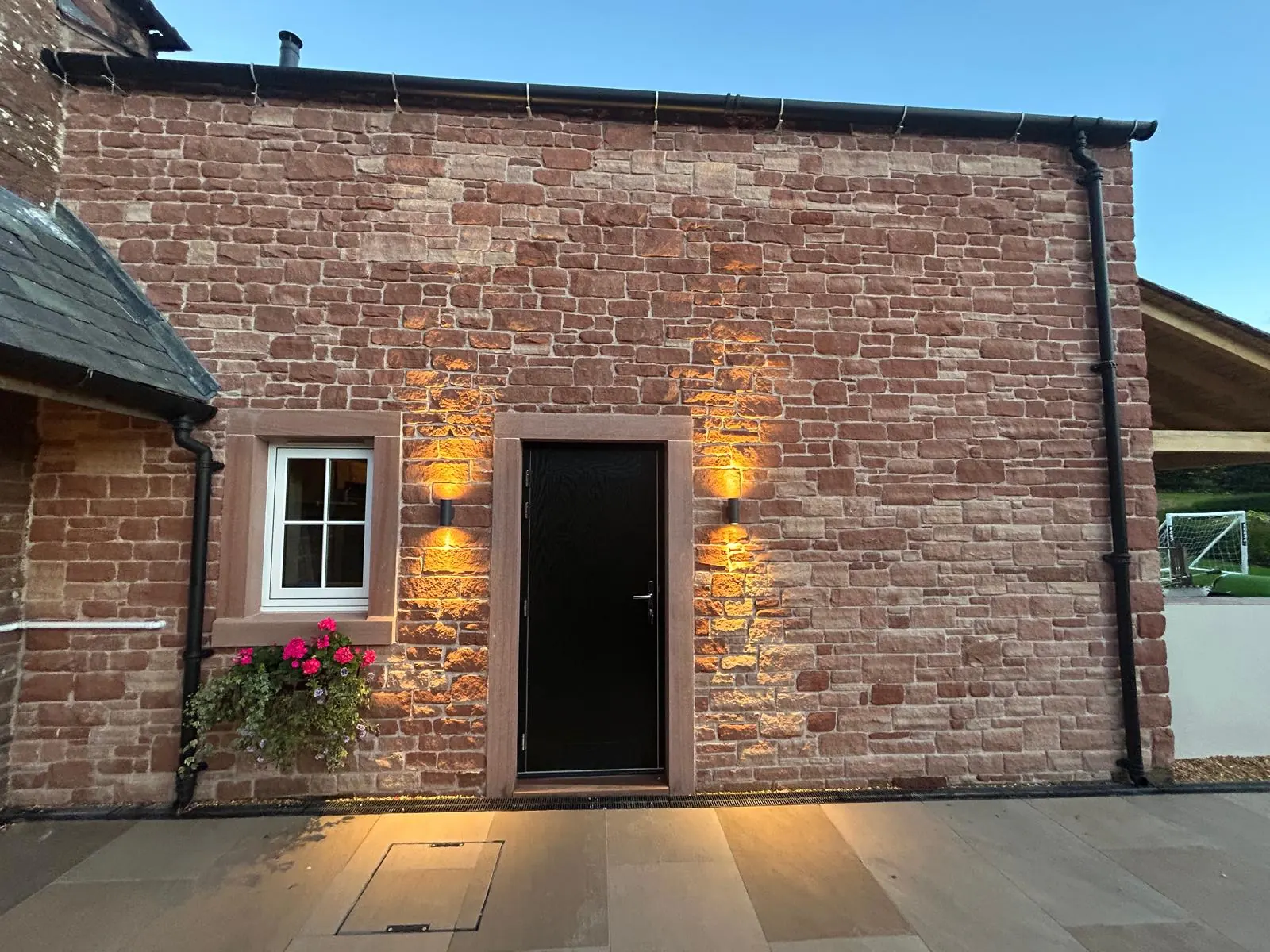 A house elevation in red sandstone at dusk with a black door and two amber wall lights